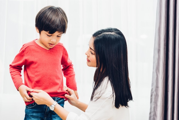 アジアの幸せな美しい母親は、学校に行く前に、家で男の子の子供に服を着せるのを手伝い、学校のコンセプトに戻る