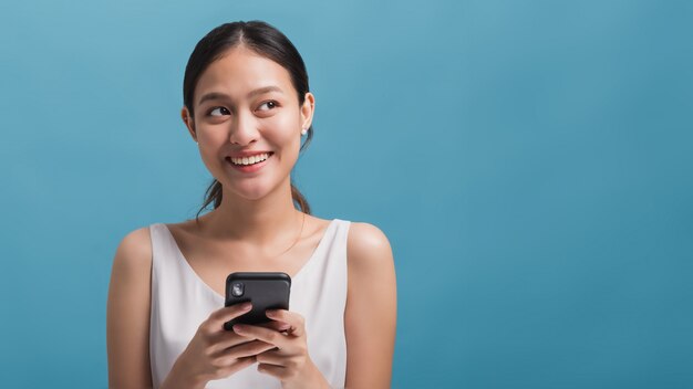 Азиатский счастливый красивый блоггер женщин усмехаясь и держа smartphone изолированный в голубой предпосылке