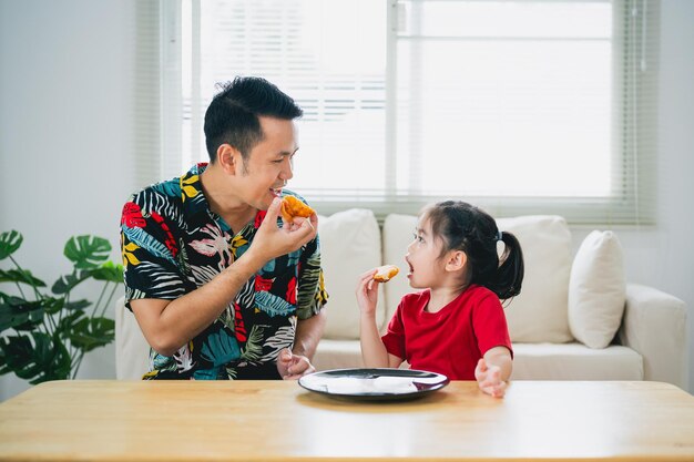 アジアの幸せな子供たちの女の子と彼女の父親は、自宅のリビングルームでピザを食べて笑顔で食事を与えます 家の中でイタリアの自家製ピザを食べて味わう子供の女の子と彼女の父親