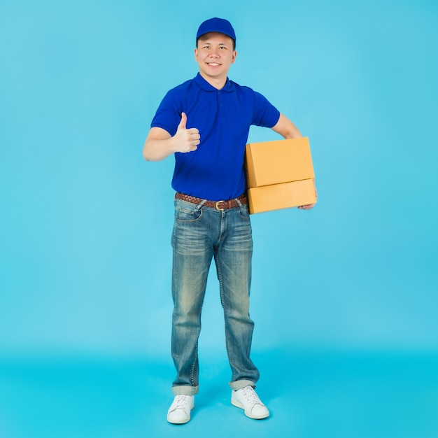 Asian happy delivery man wearing a blue shirt and hat showing thumbs up and carrying paper parcel box.