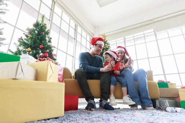 La famiglia felice asiatica e la bambina che festeggiano a natale tirano i fuochi d'artificio di carta e la scatola regalo con l'albero di natale in soggiorno. mio padre, mia madre e mia figlia con i cappelli di babbo natale seduti sul divano di casa.