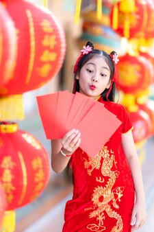 Bambina felice asiatica che indossa la decorazione tradizionale cinese cheongsam rossa che tiene in mano buste rosse e lanterne con il testo cinese benedizioni scritte su di esso è una benedizione della fortuna per il cinese