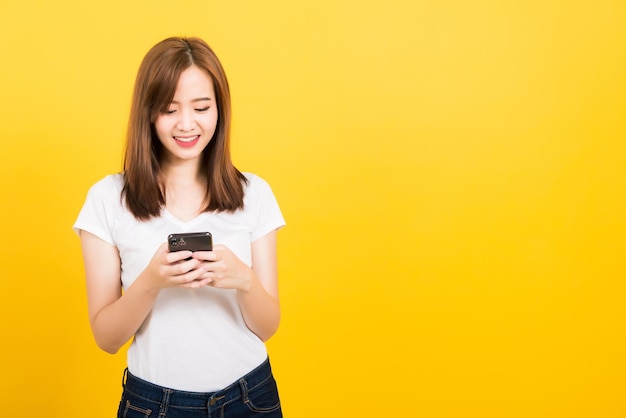 Азиатский счастливый портрет красивая милая молодая женщина-подросток улыбается стоя, играя в игру или пишу сообщение на смартфоне, глядя на изолированный телефон, студия снята на желтом фоне с копировальным пространством