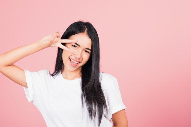 Asian happy portrait beautiful cute young woman teen smile standing showing finger making v-sign victory symbol near eye looking to camera studio shot isolated on pink background with copy space