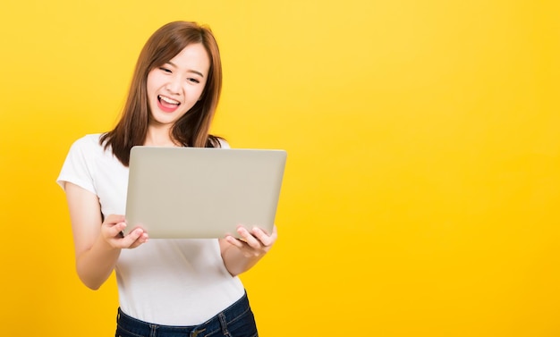 Foto ritratto felice asiatico bella bella giovane donna adolescente sorriso in piedi indossare maglietta tenere il computer portatile ed eccitato festeggiare il successo alla ricerca di camera studio sparato sullo sfondo giallo con spazio di copia