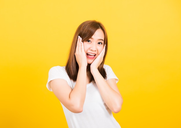 Asian happy portrait beautiful cute young woman teen stand surprised excited celebrating open mouth gesturing palms on face looking to camera isolated, studio shot yellow background with copy space