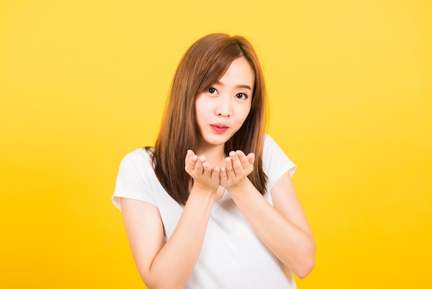 Asian happy portrait beautiful cute young woman teen standing wear t-shirt blowing kiss air something on hands looking to camera isolated, studio shot on yellow background with copy space