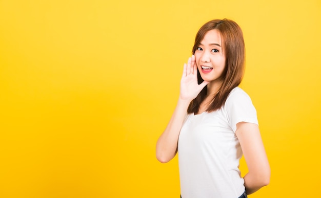 Азиатский счастливый портрет красивая милая молодая женщина-подросток стоит в футболке, рука об руку говорит, шепчет секретный слух, смотрит в камеру изолированно, студия снята на желтом фоне с копировальным пространством