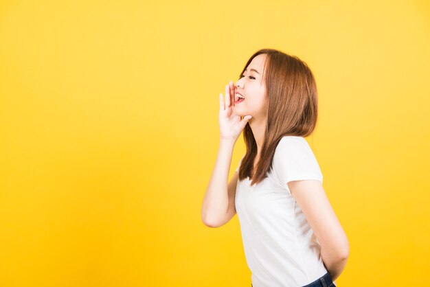 Asian happy portrait beautiful cute young woman teen standing wear t-shirt hand on mouth talking whispering secret rumor looking to side isolated, studio shot on yellow background with copy space