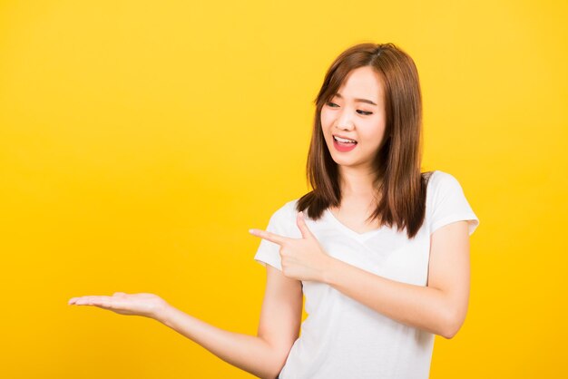 Asian happy portrait beautiful cute young woman teen standing wear t-shirt holding something on palm and point away side looking to hand isolated, studio shot on yellow background with copy space