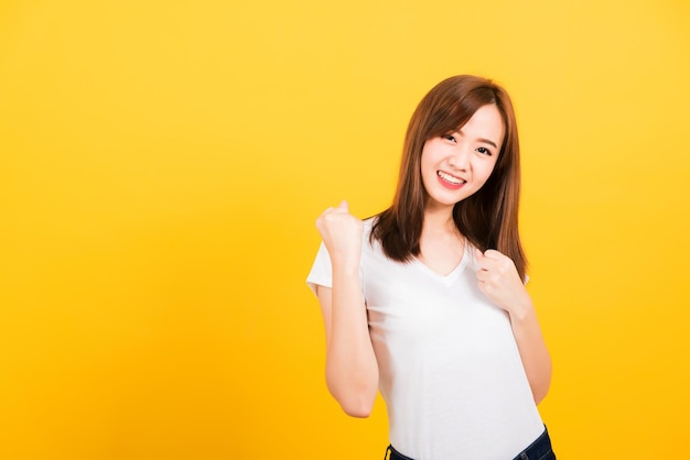 Азиатский счастливый портрет красивая симпатичная молодая женщина-подросток в стоячей одежде в футболке поднимает кулаки вверх, празднуя свой успех, глядя в камеру, изолированную, студия снята на желтом фоне с копировальным пространством