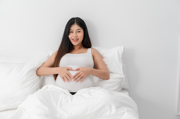 Foto la donna incinta asiatica felice è seduta sul letto e tocca la sua pancia gravidanza maternità persone e concetto di aspettativa