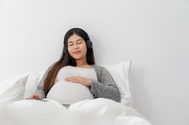아시아의 행복한 임산부는 침대에 앉아서 배를 만지고 헤드폰을 끼고 음악을 듣고 있습니다. 임신, 모성, 사람 및 기대 개념