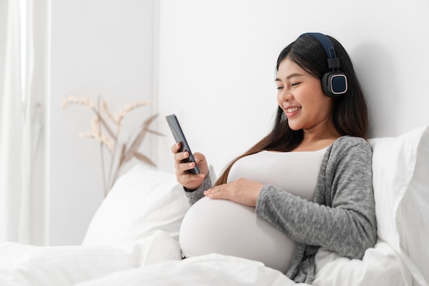 Asian happy pregnant woman is sitting on bed and and touching her belly and wear a headphone to listen the music. pregnancy, motherhood, people and expectation concept