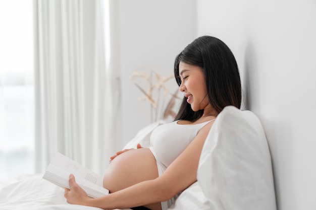 La donna incinta felice asiatica è seduta e legge un libro sul letto e tocca la sua pancia.