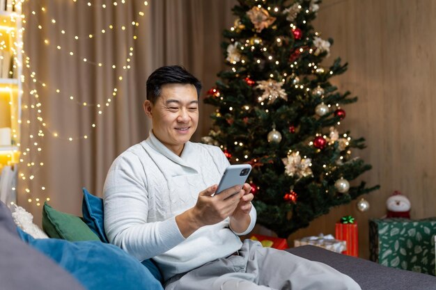 집에 있는 아시아인은 웃고 행복하게 들고 있는 전화맨을 사용하여 거실에 있는 소파에 앉아 크리스마스를 맞이합니다.