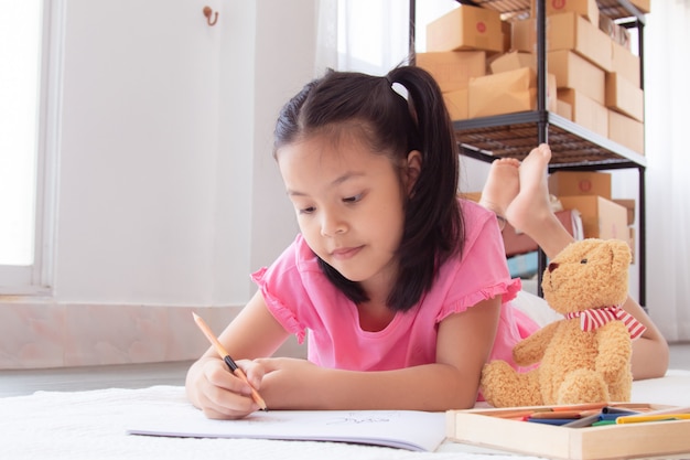 Азиатская домашняя школа. Очаровательные дети лежат на полу, рисуют домашнее задание цветными карандашами, веселая жизнерадостная девочка проводит время, концентрируясь на карантинном периоде, прежде чем вернуться в школу.
