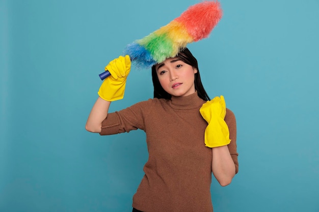 Азиатская домработница, напряженная и злая от работы по дому, держащая красочную тряпку в желтой перчатке, изолированную на синем фоне, концепция уборки дома, грустное женское выражение лица, измученная