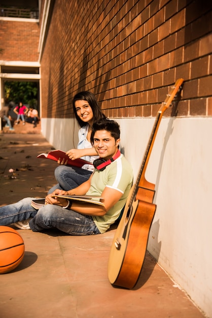 Азиатский индийский студент колледжа в фокусе работает за ноутбуком или читает книгу, в то время как другие одноклассники на заднем плане, фотография на открытом воздухе в университетском городке