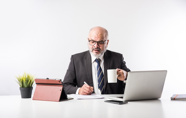 Asian Indian senior financial businessman sitting at his workstation or desk in front of a computer, laptop and tablet. Speaking on phone while doing some paperwork