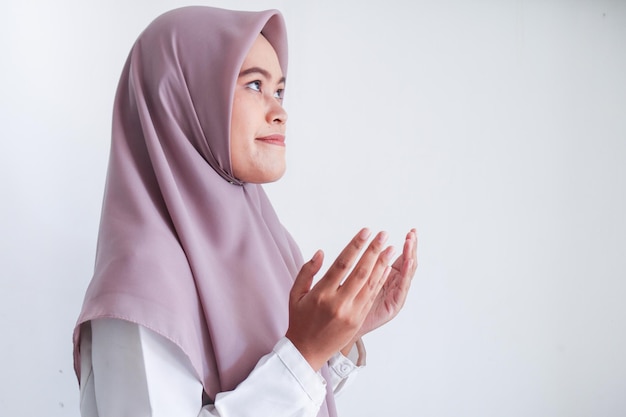 Asian islam muslim woman in headscarf and hijab prays with her\
hands up in air with smile face indonesian woman religion praying\
concept isolated on grey background