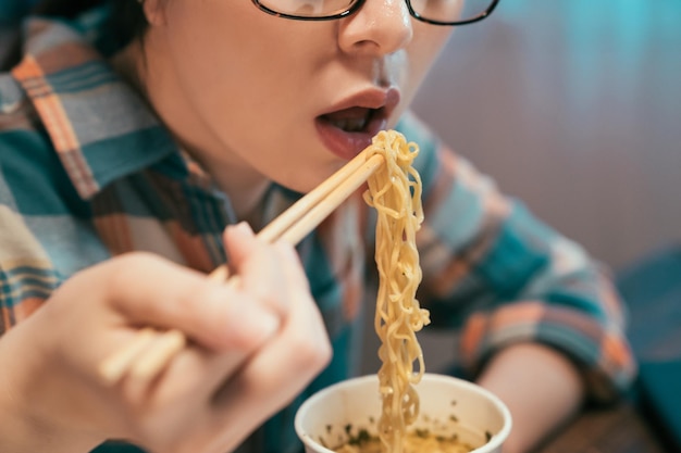 写真 アジアの日本人女性は箸でインスタントラーメンを食べます。メガネの女性をクローズアップ使い捨てカップで深夜の食事をお楽しみください。空腹の女性は口を開けて、自宅の屋内で就寝前のおやつとしてファーストフードを楽しんでいます。