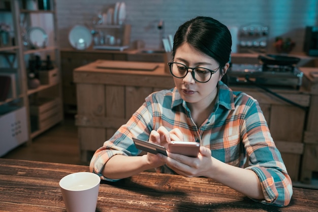 азиатская японская молодая женщина в очках делает покупки в Интернете с помощью кредитной карты, используя смартфон дома. Элегантная девушка с мобильным телефоном и дебетовой картой делает интернет-платежи на ночной кухне в доме