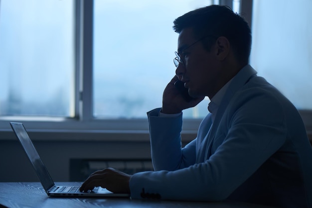 Asian Kazakh businessman in a suit and glasses with a laptop computer in the office speaks by mobile phone, solves business issues, negotiates a deal.