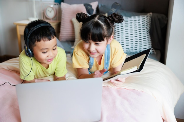 ビデオ通話の先生と一緒にオンラインで勉強するためにラップトップとヘッドフォンを使用し、かわいい妹に寝室で彼を助けてもらうアジアの少年