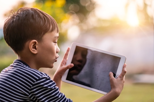 Asian kid holding playing smart tablet device playing games, sitting outdoor in garden, smiling joyfully enjoying free time, sunset shining through bokeh background, wearing short and stripy shirt