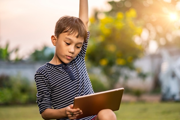 Photo asian kid holding playing on smart tablet with fist up action, sitting outside in garden, smiling joyfully enjoying free time, sunset shining through bokeh background, wearing short and stripy shirt