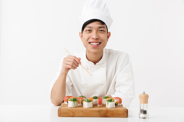 азиатский добрый главный мужчина в белой форме повара ест суши с палочками для еды, изолированными над белой стеной