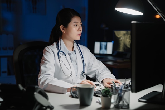 Medico femminile coreano asiatico che utilizza il computer in studio medico di notte. giovane personale ospedaliero sul posto di lavoro scuro digitando la tastiera sul record del paziente sul documento internet. operaio clinico serio e laborioso