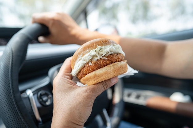 Asian lady holding hamburger to eat in car dangerous and risk an accident