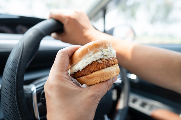 Азиатская женщина держит гамбургер, чтобы съесть в машине опасно и рисковать аварией