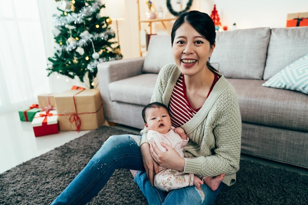 アジアの女性はカメラを見て、かわいい娘を抱き、幸せそうに輝いています。小さな赤ちゃんを腕に抱えたミレニアル世代の女性は、初めての母親であることの喜びに満ちています。