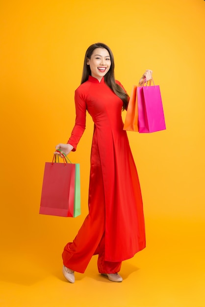 Asian lady in red ao dai dress holding shopping bags isolated on yellow, Happy lunar new year