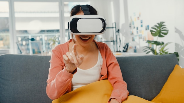 Asian lady wearing headset glasses of virtual reality\
gesticulating hand sitting on couch in living room at house.