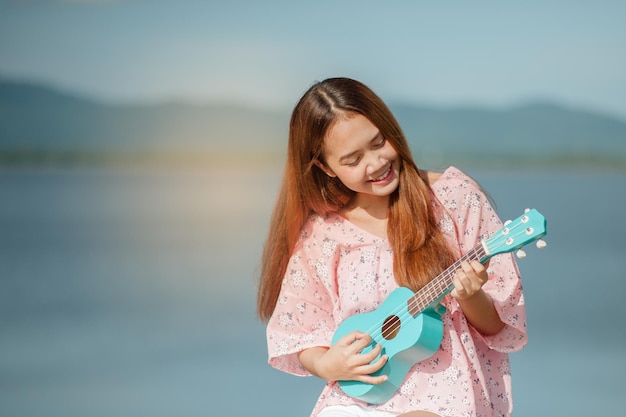 帽子をかぶったアジアの女性は、夏にウクレレのボサノバ音楽を演奏します屋外で音楽を演奏する若いかわいい女性幸せな若い女の子は、湖の下でウクレレを楽しんでいます