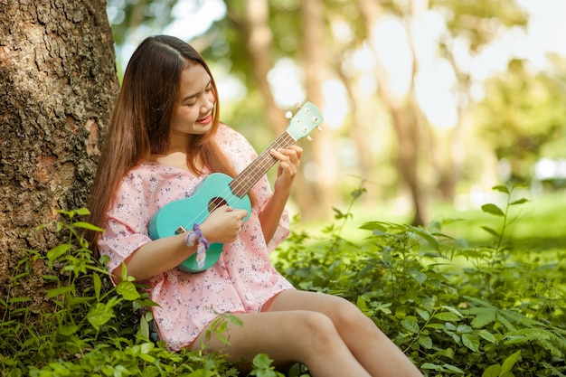 Asian lady with hat play ukulele bossanova music in summer timeYoung cute woman playing music outdoorsA happy young girl enjoys playing ukulele under a tree Beautiful nature in the background