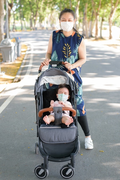 庭でマスクを身に着けている母親と一緒にベビーカーに座っているフェイスマスクを持つアジアの小さな女の赤ちゃん