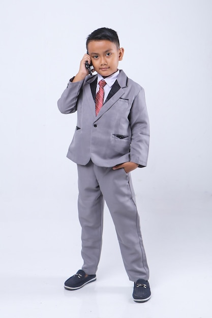 Asian little boy in formalwear talking on the mobile phone while standing and look at camera.