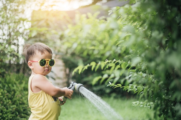 アジアの少年は木に水を注いでいます。子供は庭でじょうろで植物の世話をするのに役立ちます。