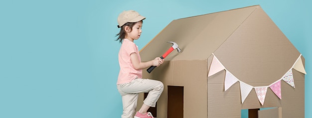 Азиатская девочка маленького ребенка, строящая ее картонный дом, изолированный на синем длинном баннере с копией, делает интервалы для вашего текста, творческого дома с концепцией семьи