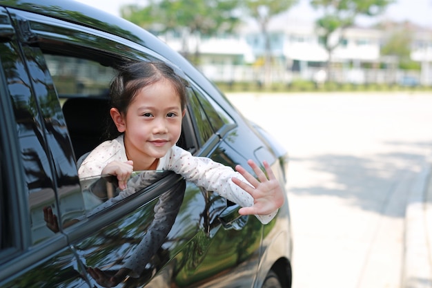 車の中でアジアの小さな子供の女の子が笑顔でカメラを見て、車の座席に座ってさよならを振っています。