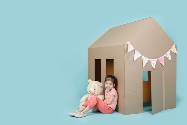 Asian little child girl playing with cardboard house with her teddy bear isolated on blue long banner with copy space for your text, Creative at home with family concept