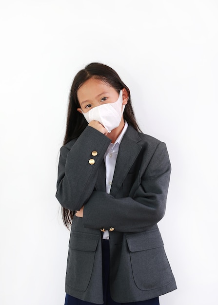 Азиатская маленькая девочка носит формальную рубашку, студенческий костюм и защитную маску для лица и держит руки скрещенными на белом фоне