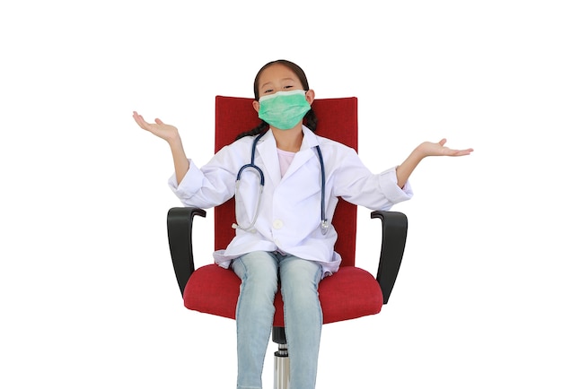 Азиатский ребенок маленькой девочки в белом халате доктора и защитной маске с широко распростертыми объятиями сидя на красном стуле изолированном на белой предпосылке студии. Изображение с обтравочным контуром