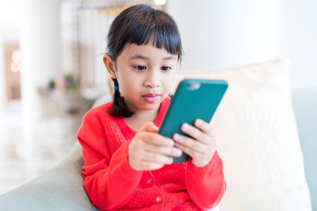 アジアの小さな女の子が家で携帯電話で遊んでいます