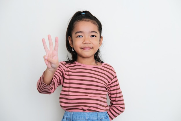 Азиатская маленькая девочка улыбается в камеру и показывает знак тремя пальцами